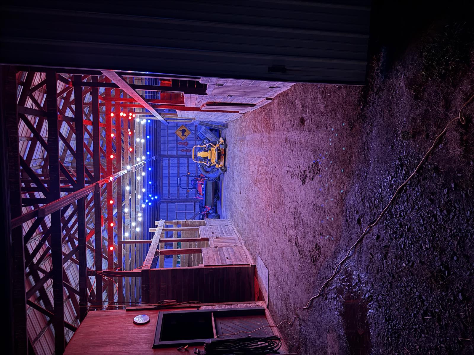 Inside the barn at night on the Fourth of July, red, white, and blue string lights along the rafters, a tractor and a mower parked at the far end, an extension cord running across the dirt floor