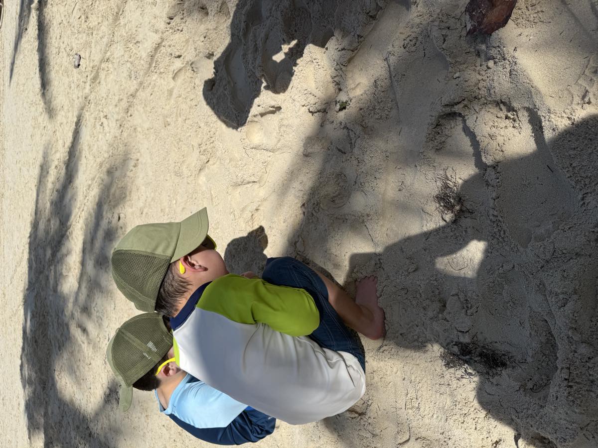 Two boys in caps crouched over a hole they are digging in the sand, photographed from behind