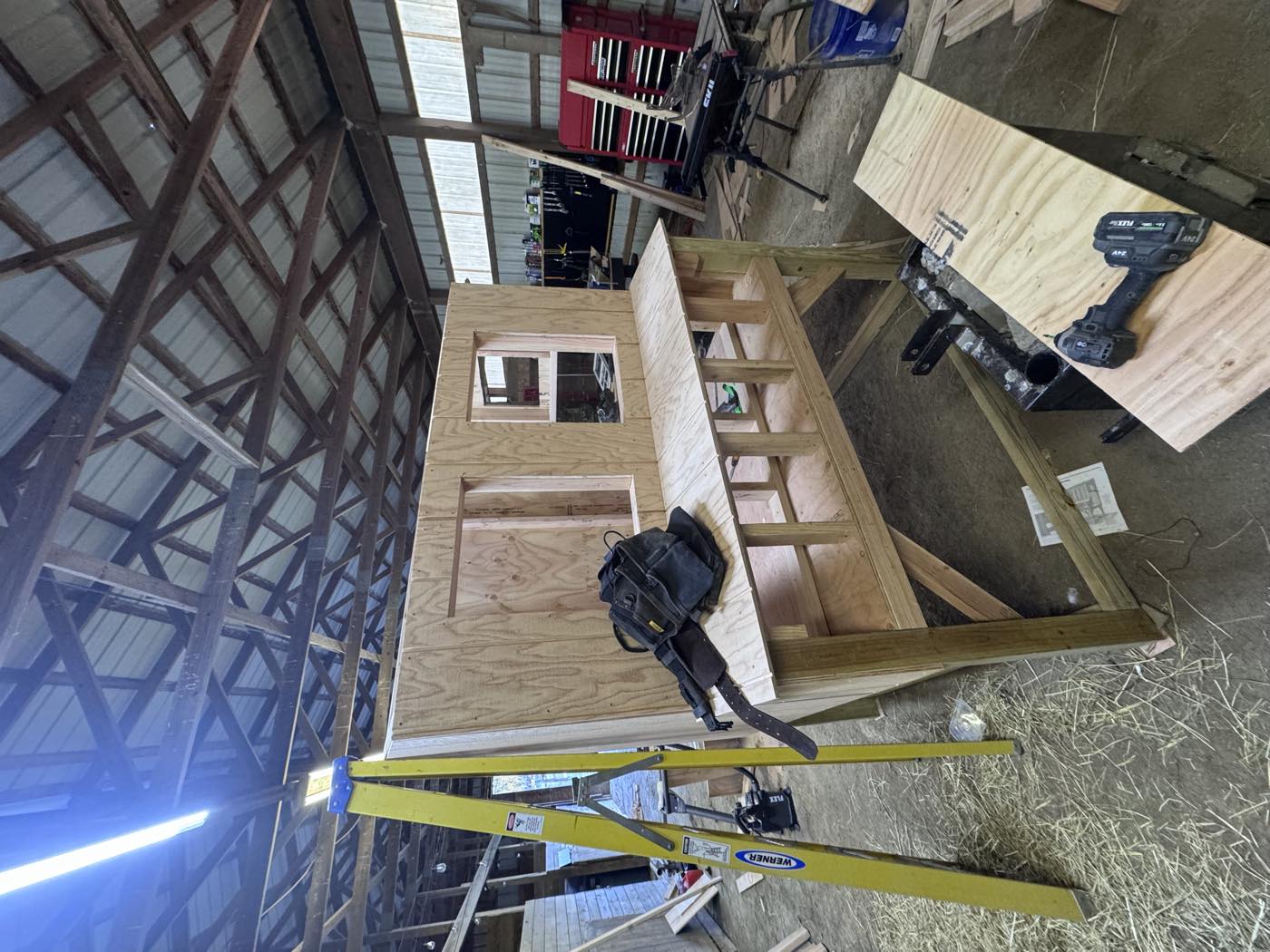 The coop frame mid-build on a workbench inside the shop, plywood walls up, tool bag resting on the deck, ladder alongside