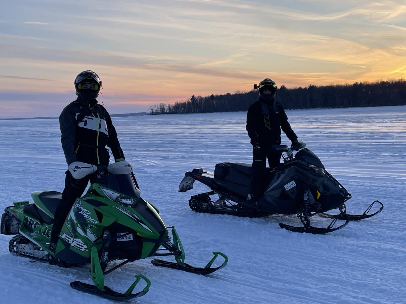 Two snowmobiles stopped on a frozen lake at sunset, riders in helmets, treeline on the horizon