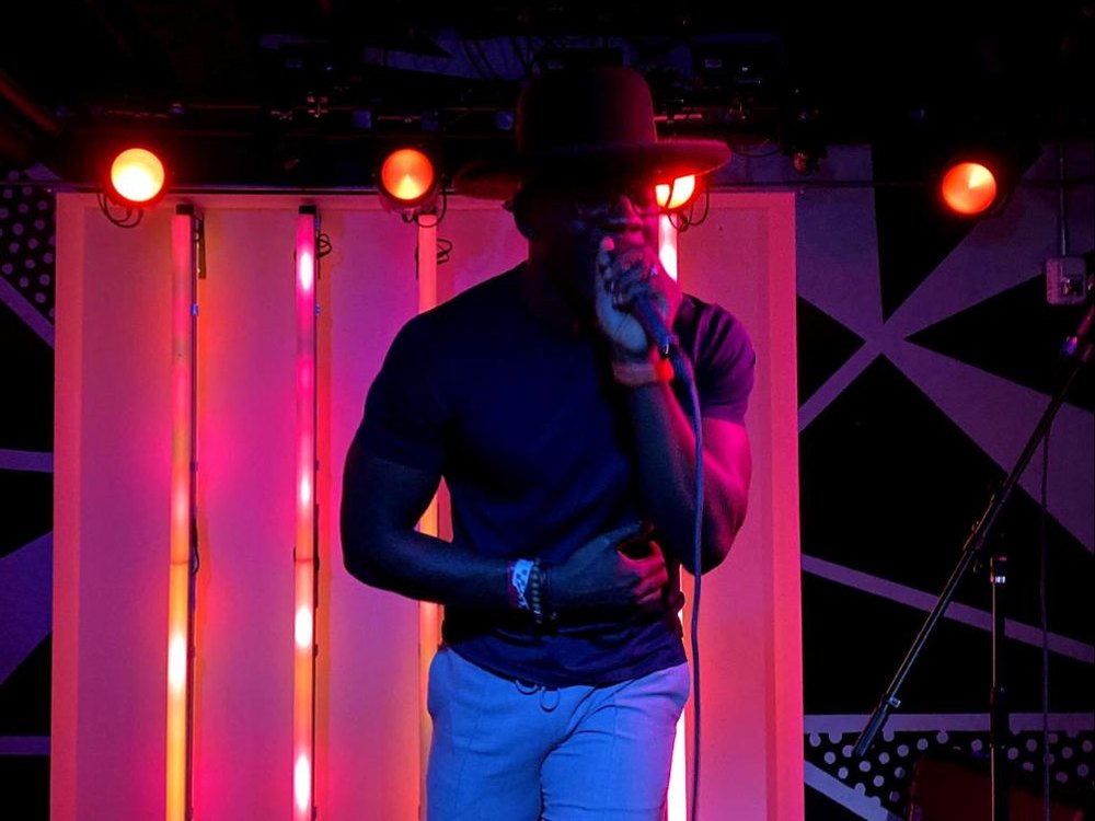 A performer at the microphone on the summit's late-show stage, hat pulled low, under pink and orange stage lights